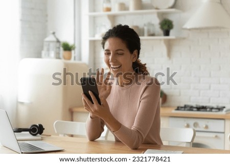 Joyful young hispanic caucasian woman looking at cellphone screen, enjoying distant web camera call using mobile video call zoom application, involved in pleasant distant conversation in kitchen. Royalty-Free Stock Photo #2321778421