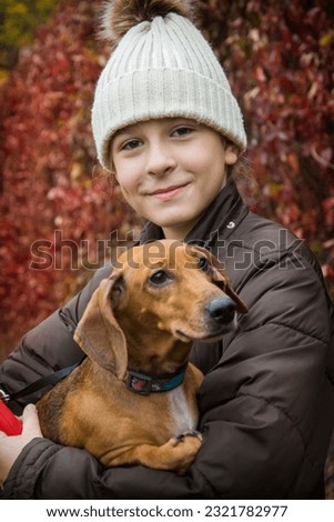 In the autumn park, a girl holds a dachshund dog in her arms.