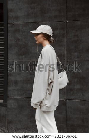 portrait of beautiful blonde girl dressed in beige oversized sweatshirt, white sweatpants, cap, bag, phone hangs over shoulder, stylish fashion sports outfit, lifestyle model Royalty-Free Stock Photo #2321786761