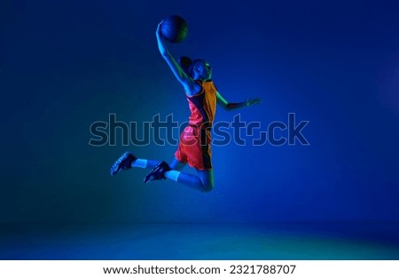 Slam dunk. Motivated young girl, basketball player throwing ball in jump against blue studio background in neon light. Concept of professional sport, action and motion, game, competition, hobby, ad