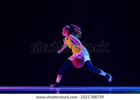 Young female athlete, professional basketball player in motion, dribbling ball over black studio background in neon light. Professional sport, action and motion, game, competition, hobby, ad concept Royalty-Free Stock Photo #2321788759