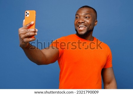 Young man of African American ethnicity he wear orange t-shirt doing selfie shot on mobile cell phone post photo on social network isolated on plain dark royal navy blue background. Lifestyle concept