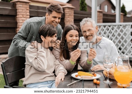 Smiling teen girl using smartphone near middle aged parents and brother during barbeque party with summer food and parents day celebration at backyard in june, happy parents day concept Royalty-Free Stock Photo #2321794451