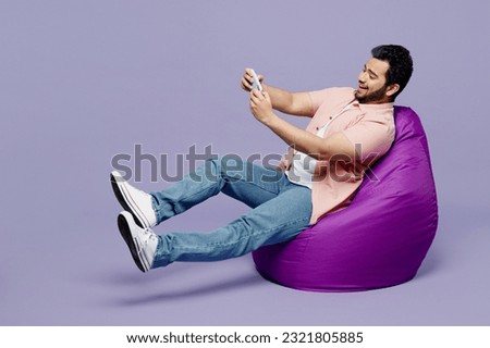 Full body young man wearing pink shirt white t-shirt casual clothes sit in bag chair use play racing app on mobile cell phone gadget smartphone for pc video games isolated on plain purple background