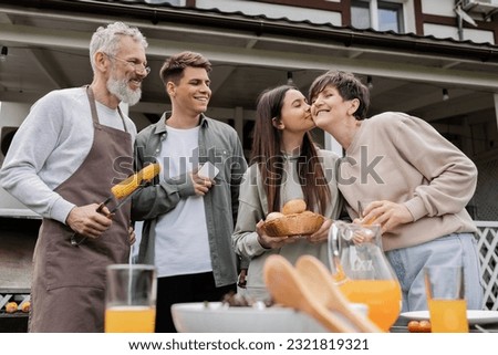 parents day, happy middle aged parents, having family bbq party, teenage daughter kissing cheek of mother, siblings, father holding tongs with grilled corn, summer house, suburban life