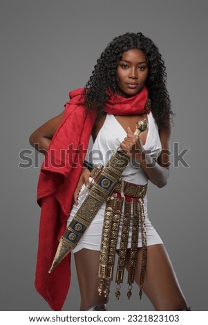 Stunning dark-skinned beauty dons a white Greek tunic with a striking red cape, intricately decorated belt and greaves and confidently wields richly adorned gladius sword on grey background