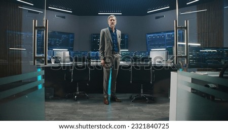Mature big data scientist in business suit puts on glasses, stands in monitoring room and looks at camera. Multiple big screens on the wall with displayed live analysis feed at background. Dolly shot.