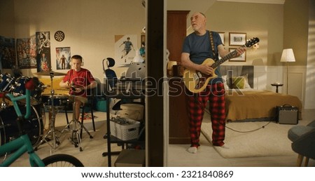 Young boy practices musician skills uses drum kit. Elderly man sings and plays on electric guitar with neighbour. Thin walls and low level of sound insulation. Neighbors play music together. Royalty-Free Stock Photo #2321840869