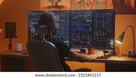 Male stock trader analyzes real-time stocks, exchange market charts on multi-monitor computer workstation, eats french fries. Man works in investment at home office at night. Cryptocurrency trading.