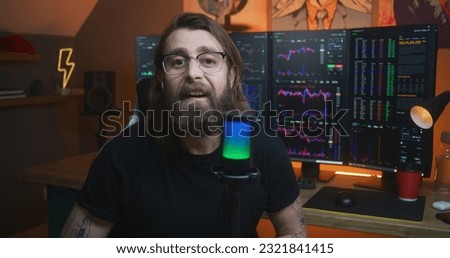 Male stock trader looks at camera and speaks into microphone. Man records educational video training for blog about investment. Multi-monitor computer with real-time stocks, exchange market charts.
