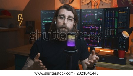 Male stock trader looks at camera and speaks into microphone. Man records educational video training for blog about investment. Multi-monitor computer with real-time stocks, exchange market charts.