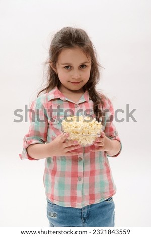 Isolated portrait on white background of beautiful lovely Caucasian little child girl in blue denim jeans and checkered shirt, holding a bowl of delicious pop corn and cutely smiling looking at camera