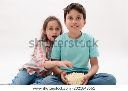 Two amazed adorable diverse kids, teenage boy and little girl, brother and sister sitting over white isolated studio background, watching movie, eating popcorn, expressing wow emotion and excitement
