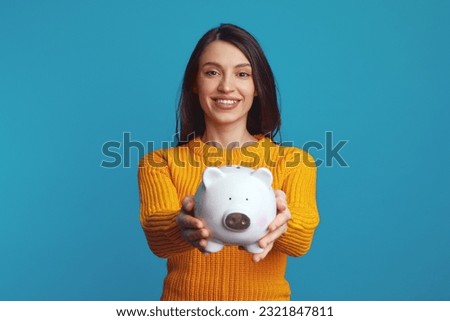 Cheerful young girl in casual orange sweater smiling and holding white piggy bank with lots of money isolated over blue background