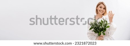happiness, joyful bride with blonde hair standing in white silk robe and holding bridal bouquet, showing engagement ring, young woman, beautiful, excitement, feminine, white flowers, banner Royalty-Free Stock Photo #2321872023