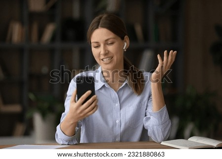 Beautiful happy young business lady in wireless earphones looking at mobile phone screen, holding online video call web camera negotiations meeting with colleagues, discussing working issues distantly Royalty-Free Stock Photo #2321880863