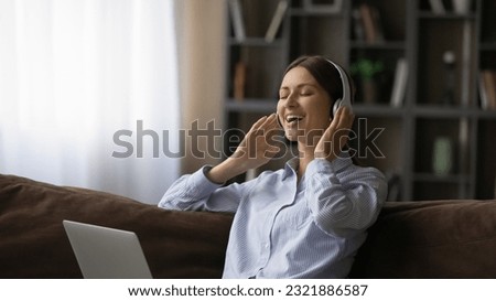 Happy relaxed attractive young woman listening favorite music in wireless headphones, enjoying stress free leisure weekend time on couch with computer on laps, domestic hobby activity concept. Royalty-Free Stock Photo #2321886587