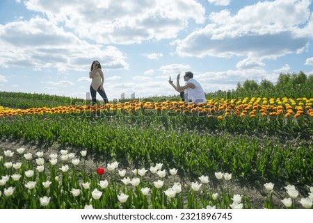 A guy takes a picture of his girlfriend with his phone at field of colorful tulips background. Young beautiful couple in sport clothes, on the background of a field of colorful tulips.