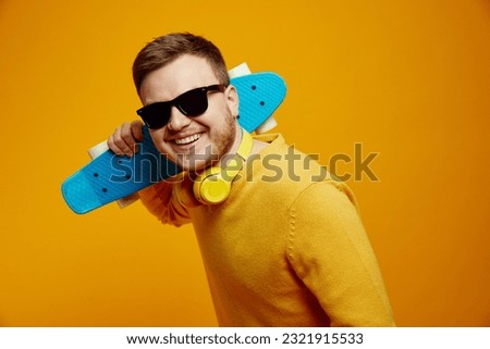 Cheerful cool guy wearing yellow jumper and trendy sunglasses holding longboard on shoulders looking super excited, isolated over orange background Royalty-Free Stock Photo #2321915533
