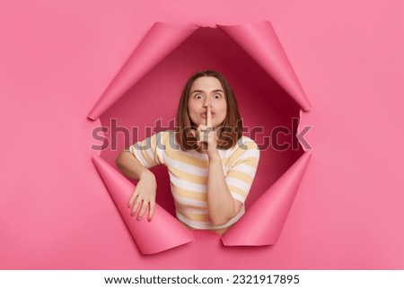 Beautiful brunette woman wearing striped top looking through ripped pink paper hole keeps finger near lips keeps secret showing shh gesture to camera.