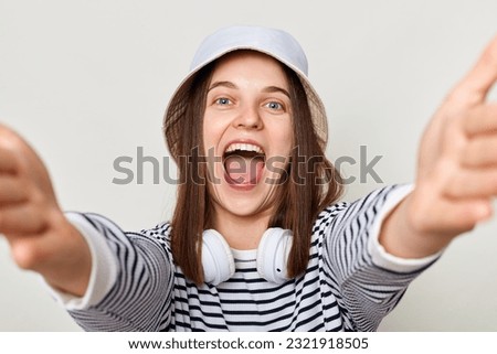 Joyful extremely happy woman wearing striped shirt and panama standing with headphones over neck isolated over gray background making point of view photo screaming with happiness.