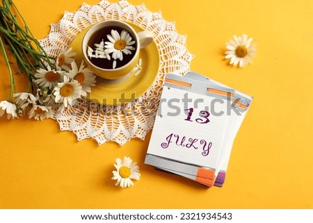 Calendar for July 13: the name of the month July in English, the numbers 13, a cup of tea on an openwork napkin with chamomile, a bouquet of daisies next to it, yellow background