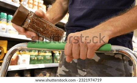 Close-up of a shopping cart in a supermarket and a male buyer's hand putting a glass jar of instant coffee into it Royalty-Free Stock Photo #2321974719