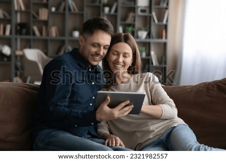 Smiling millennial man and woman relax on sofa in living room use modern tablet gadget together. Happy young Caucasian couple rest on couch at home speak talk on webcam video call on pad.