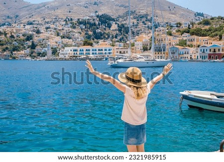 Cute blonde girl child on holiday in Greece. Symi also known as Syme or Simi, Greek island on the background. Symi main city with colorful buildings on background. Dodecanese islands.