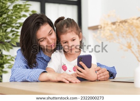 Head shot smiling young mother showing funny cartoons to overjoyed little adorable girl on smartphone. Happy 30s woman recording family video 