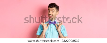 Classy handsome man fixing bow-tie on neck and smiling, getting ready for date or party, standing on pink background. Royalty-Free Stock Photo #2322008537