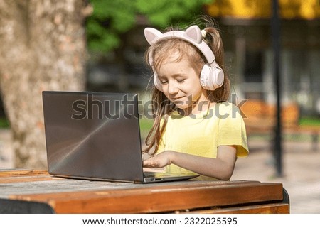 Little girl with pink headphones, wearing a yellow t-shirt, sitting in front of the laptop, typing and moving the mouse, on the terrace. Online distance education, childhood and technology concept.