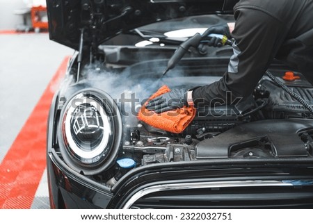 Car detailing concept. Manual engine cleaning with steam. Car detailing expert deep-cleaning black vehicle and wiping car surface with orange microfibre cloth. High quality photo