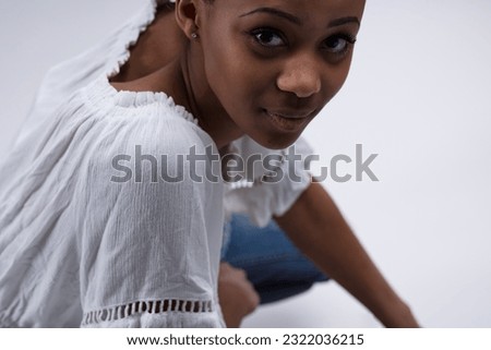 Portrait from below of a young woman sitting, turning to listen. Wearing a white blouse and jeans, she seems curious if you're interesting or dull. She's black and very pretty Royalty-Free Stock Photo #2322036215