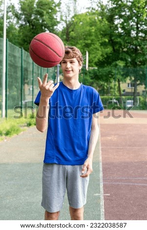 Cute boy in t shirt plays basketball on city playground. Active teen enjoying outdoor game with red ball. Hobby, active lifestyle, sport for kids.