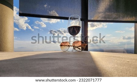 A glass of red wine and sunglasses on the corner of a table with a white tablecloth, office buildings and skyscrapers in the city skyline at sunset with blue sky and clouds at The Sun Dial Restaurant