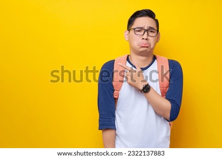 Sad and unhappy young Asian man student in casual clothes bag glasses pointing finger at copy space and frowning isolated on yellow background. Education in high school university college concept Royalty-Free Stock Photo #2322137883
