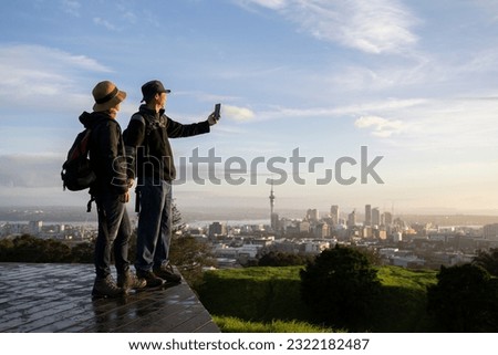 Couple taking selfie with smartphone on Mt Eden summit at sunrise. Skytower in the background. Selective focus on people in foreground. Auckland. 