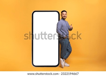 Mobile Offer. Excited asian guy leaning on big smartphone with blank screen and showing thumb up gesture, millennial man recommending great new app or website, yellow background, mockup Royalty-Free Stock Photo #2322194667
