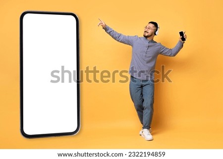 Mobile App. Happy Asian Man Wearing Headphones Dancing Near Big Blank Smartphone With White Screen, Cheerful Millennial Guy Having Fin Over Yellow Studio Background, Collage, Mockup