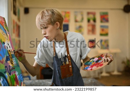 Concentrated teenage asian gay man dressed in apron painting picture with oil paints on canvas in bright art class Royalty-Free Stock Photo #2322199857