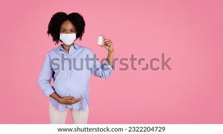 Smiling millennial african american curly pregnant woman in casual, protective mask touch big belly with hand, show pills jar, isolated on pink studio background. Health care, vitamins and minerals