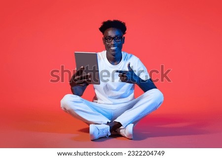 Cheerful handsome cool young black guy wearing white casual outfit and eyeglasses sitting on floor, using modern gadget digital tablet on red background in neon light, recommending nice app
