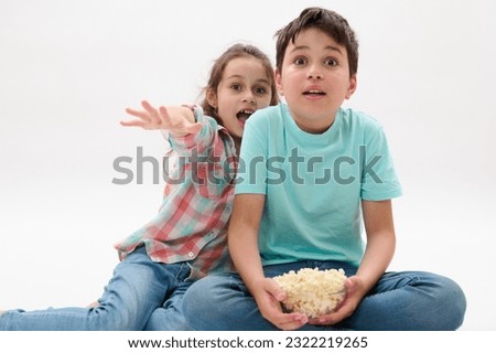 Adorable two diverse children with bowl of popcorn, looking toward the camera with frightened look, watching scary or fantasy movie, expressing fright and fear, isolated over white studio background