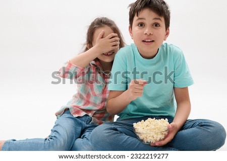 Amazed kids, teen boy and little child girl covering her eyes from fright, watching scary movie or cartoon, eating popcorn over white isolated background. People. Kids. Leisure activity. Entertainment