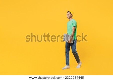 Full body fun young IT man of African American ethnicity he wears casual clothes green t-shirt hat hold closed laptop pc computer isolated on plain yellow background studio portrait Lifestyle concept