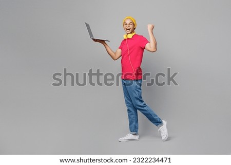 Full body fun side view young man of African American ethnicity wear pink t-shirt yellow hat headphones hold use work on laptop pc computer do winner gesture isolated on plain grey background studio Royalty-Free Stock Photo #2322234471