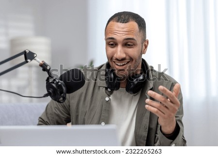 Close-up photo. Young hispanic man sitting at home wearing headphones, talking into a microphone on a video call from a laptop. He looks smilingly at the monitor, communicates.