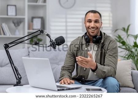 Portrait of a young African-American man sitting at home on the sofa in front of a microphone and a laptop. He looks at the camera with a smile. Records video, audio, blogs, podcasts, radio host.