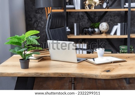 Fragment of the office. Desk with notebook, notepad, pen and plant. Nearby is a beautiful black leather armchair. In the background are shelves with books, globes, clocks and other decor.
